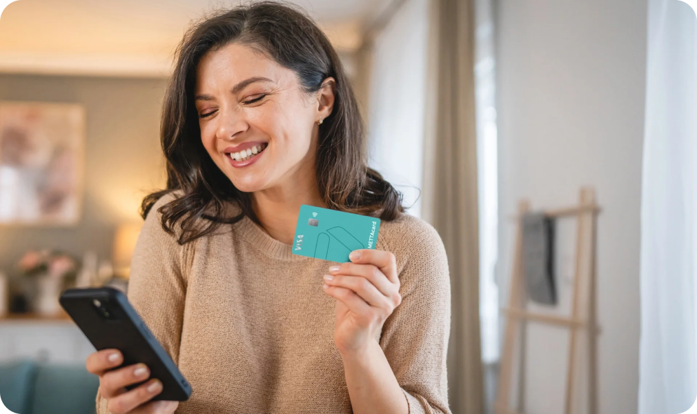 Mulher sorridente com cartão METTAcard na mão e com o celular em outra mão
