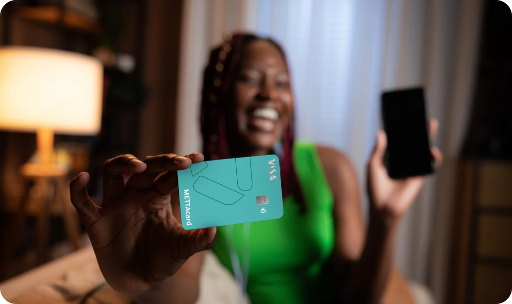 Mulher sorridente com cartão METTAcard na mão e com o celular em outra mão